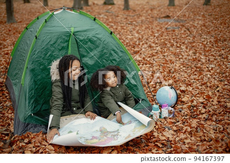 Beautiful little black girls in tent camping in the forest. Two little sisters sitting in a tent in autumn forest and looking on a map. Black girls wearing khaki coats. Beautiful little black girls in tent camping in the forest. Two little sisters sitting in a tent in autumn forest and looking on a map. Black girls wearing khaki coats. 94167397