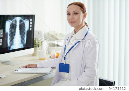 Portrait of young female pulmonologist checking x-ray of recovering patient and filling document 94173212