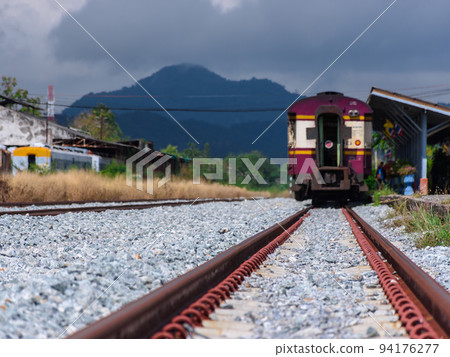 Ground level view of the railway at the train station in countryside with a train, mountain and cloudy sky as a blurry background. 94176277