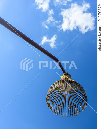 Bottom-up view of old street lamp made from bamboo designed as chicken coop. It is hung on brown pillar made from wood and metal in public place with beautiful blue sky and white cloud as background. Bottom-up view of old street lamp made from bamboo designed as chicken coop. It is hung on brown pillar made from wood and metal in public place with beautiful blue sky and white cloud as background. 94176279