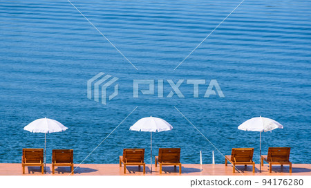 Beautiful view of white umbrellas and wooden chairs placed on wooden ground that is floating over the public lake. Complementary color. Beautiful view of white umbrellas and wooden chairs placed on wooden ground that is floating over the public lake. Complementary color. 94176280