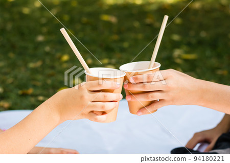 Two children's hands are holding eco-friendly cups and tubes with apple juice at a picnic in the park. Eco-friendly disposable tableware 94180221