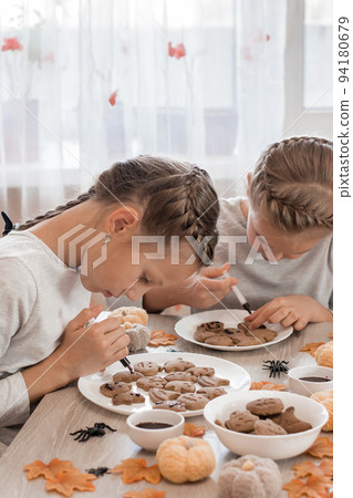 Preparing to celebrate halloween and preparing a treat. Two girls decorate halloween gingerbread cookies on plates with chocolate icing. Lifestyle 94180679