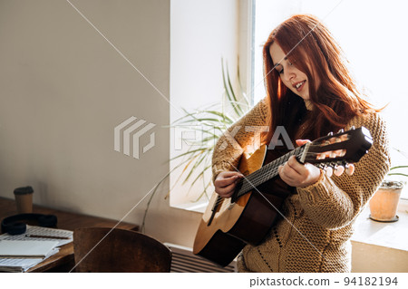 Indoor cozy Hobbies for winter, autumn cold season. Redhead woman playing acoustic guitar and listening to music, sitting near window home Indoor cozy Hobbies for winter, autumn cold season. Redhead woman playing acoustic guitar and listening to music, sitting near window home 94182194
