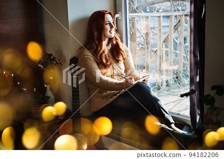 Indoor cozy Hobbies for winter, autumn cold season. Redhead woman playing acoustic guitar and listening to music, sitting near window home 94182206