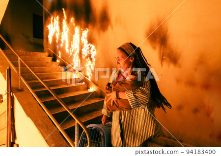 Brave Fireman Descends Stairs of a Burning Building and Holds Saved Girl in His Arms. Open fire and one Firefighter in the Background. 94183400