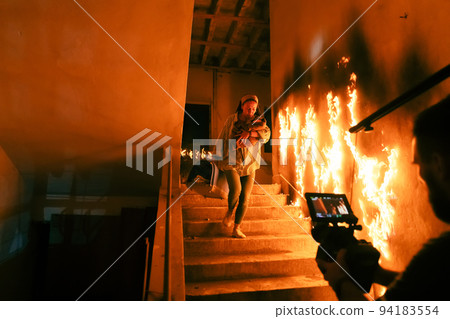 Brave Fireman Descends Stairs of a Burning Building and Holds Saved Girl in His Arms. Open fire and one Firefighter in the Background. 94183554