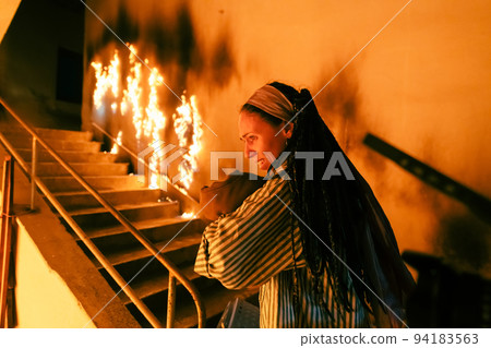 Brave Fireman Descends Stairs of a Burning Building and Holds Saved Girl in His Arms. Open fire and one Firefighter in the Background. 94183563