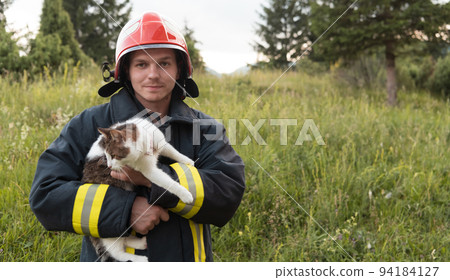 Close-up portrait of heroic fireman in protective suit and red helmet holds saved cat in his arms. Firefighter in fire fighting operation. 94184127