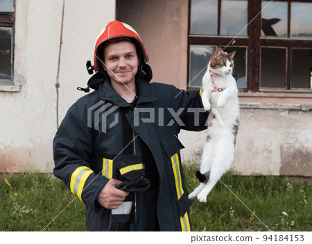 Close-up portrait of heroic fireman in protective suit and red helmet holds saved cat in his arms. Firefighter in fire fighting operation. Close-up portrait of heroic fireman in protective suit and red helmet holds saved cat in his arms. Firefighter in fire fighting operation. 94184153
