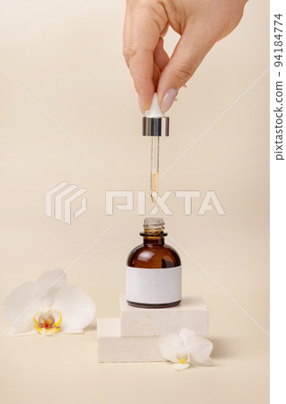 Hand with pipette over opened cosmetic dropper bottle near white orchid flowers, Mockup 94184774