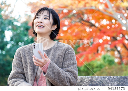 A young woman using a smartphone in a park with autumn leaves A young woman using a smartphone in a park with autumn leaves 94185373