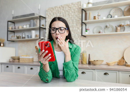 A young woman looks shocked and surprised at the phone, covered her mouth with her hand, saw a message from her ex-lover, learned about betrayal. Sitting at home in the kitchen and holding a red phone A young woman looks shocked and surprised at the phone, covered her mouth with her hand, saw a message from her ex-lover, learned about betrayal. Sitting at home in the kitchen and holding a red phone 94186054