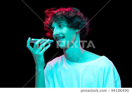 Portrait of young man with mustache recording voice message on phone isolated over black background in neon light 94186490