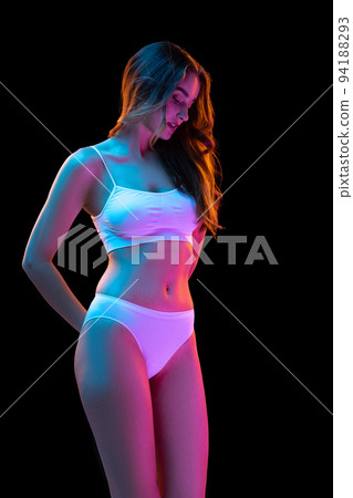 Studio shot of young charming slim girl in white cotton underwear isolated over dark background in neon light. Wellness, fitness, diet, fashion, natural beauty Studio shot of young charming slim girl in white cotton underwear isolated over dark background in neon light. Wellness, fitness, diet, fashion, natural beauty 94188293