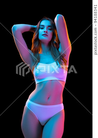 Studio shot of young charming slim girl in white cotton underwear isolated over dark background in neon light. Wellness, fitness, diet, fashion, natural beauty Studio shot of young charming slim girl in white cotton underwear isolated over dark background in neon light. Wellness, fitness, diet, fashion, natural beauty 94188341