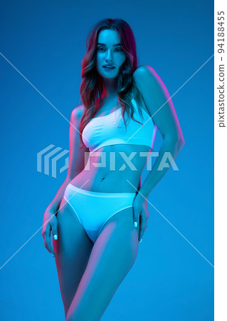 Portrait of young sensual woman in white cotton underwear isolated over blue background in neon light. Wellness, fitness, diet, beauty of female body 94188455
