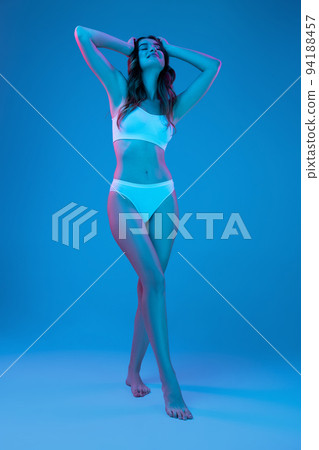 Studio shot of young charming slim girl in white cotton underwear isolated over blue background in neon light. Wellness, fitness, diet, fashion, natural beauty 94188457