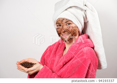 A woman in a pink bathrobe with a coffee mask on her face holds coffee beans in her hands 94193337