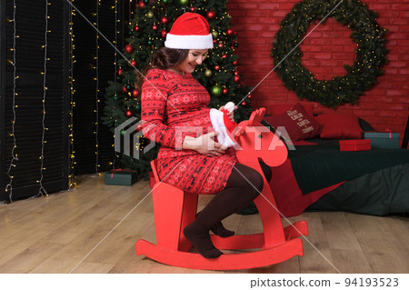 A pregnant woman prepares for Christmas, sits on a red children's wooden horse and puts a hat on her belly. A pregnant woman prepares for Christmas, sits on a red children's wooden horse and puts a hat on her belly. 94193523