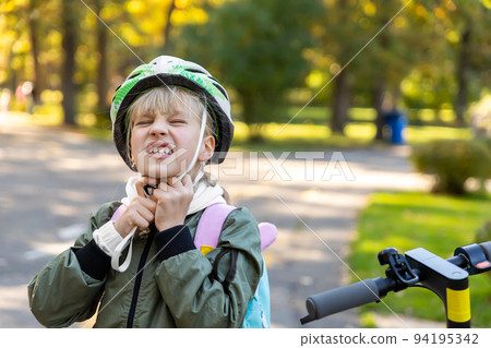 Profile view portrait cute blond little caucasian school girl putting on wrong size painful helmet riding electric scooter city street park outdoors sunny day. Healthy sport children activity outside 94195342