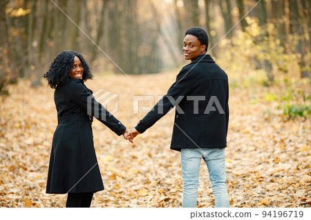 Black young man and his girlfriend holding hands. Romantic couple walking in autumn park on sunset. Man and woman wearing black coats. 94196719