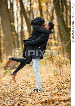 Black young man holding his girlfriend on hands. Romantic couple walking in autumn park. Man and woman wearing black coats. Black young man holding his girlfriend on hands. Romantic couple walking in autumn park. Man and woman wearing black coats. 94196739