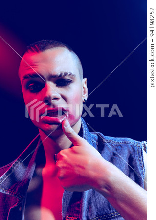 Close-up portrait of young man wearing makeup, smokey eyes, posing isolated over dark background in neon light. Informal Close-up portrait of young man wearing makeup, smokey eyes, posing isolated over dark background in neon light. Informal 94198252