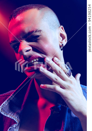 Close-up portrait of young man wearing makeup, smokey eyes, posing isolated over blue background in neon light. Showing teeth 94198254