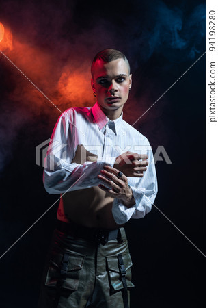 Portrait of stylish man in makeup posing in short white shirt and leather shorts over black background in neon light. Blushing cheeks Portrait of stylish man in makeup posing in short white shirt and leather shorts over black background in neon light. Blushing cheeks 94198280