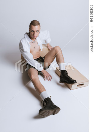 Portrait of young man in makeup, smoky eyes, posing in short shirt and leather shorts isolated over grey studio background Portrait of young man in makeup, smoky eyes, posing in short shirt and leather shorts isolated over grey studio background 94198302