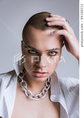 Portrait of young man with smoky eyes makeup and partly shaved brows posing over grey studio background. Big silver chain necklace Portrait of young man with smoky eyes makeup and partly shaved brows posing over grey studio background. Big silver chain necklace 94198313