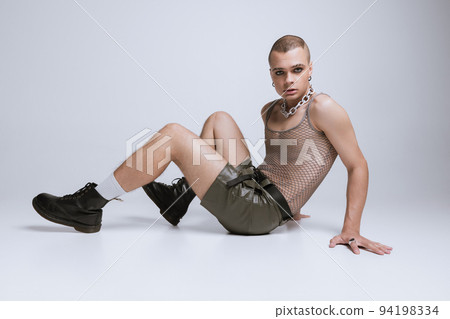Portrait of young man in makeup, smoky eyes, posing in string vest and shorts isolated over grey studio background 94198334