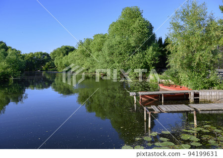 A river with water lilies, wooden piers and a boat surrounded by trees in summer A river with water lilies, wooden piers and a boat surrounded by trees in summer 94199631