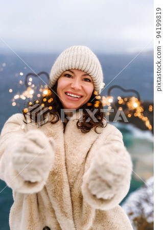 Outdoor winter portrait of elegant happy smiling middle aged woman in beige hat, light faux fur coat holding a sparkler, posing against the background of the sea and snow. Outdoor winter portrait of elegant happy smiling middle aged woman in beige hat, light faux fur coat holding a sparkler, posing against the background of the sea and snow. 94199819