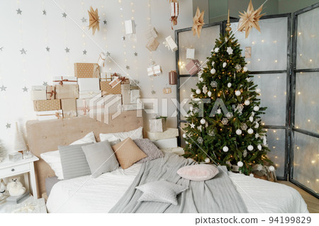 Pile of Christmas gifts in colorful wrapping with ribbons against a white wall with silver stars. Christmas tree in the room. Pile of Christmas gifts in colorful wrapping with ribbons against a white wall with silver stars. Christmas tree in the room. 94199829
