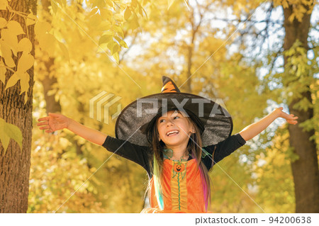 A happy girl dressed up as witch laughs from the top of mouth looking away with arms outstretched against the background of autumn park with yellow foliage. Halloween Holiday A happy girl dressed up as witch laughs from the top of mouth looking away with arms outstretched against the background of autumn park with yellow foliage. Halloween Holiday 94200638