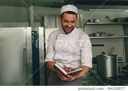 Smiling chef in uniform making notes in notebook standing on kitchen Smiling chef in uniform making notes in notebook standing on kitchen 94200877