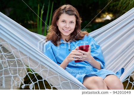 Outdoor portrait of teenage girl sitting in hammock with smartphone Outdoor portrait of teenage girl sitting in hammock with smartphone 94201049