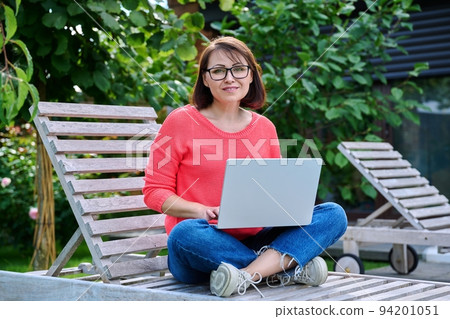 Mature woman sitting in wooden deck chair using laptop for leisure and work 94201051
