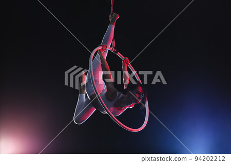 Young woman performing acrobatic element on aerial ring against dark background Young woman performing acrobatic element on aerial ring against dark background 94202212
