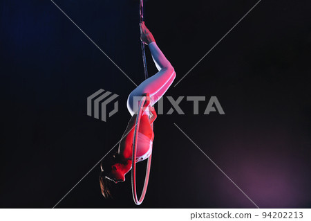 Young woman performing acrobatic element on aerial ring against dark background Young woman performing acrobatic element on aerial ring against dark background 94202213