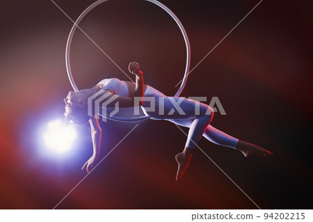 Young woman performing acrobatic element on aerial ring against dark background Young woman performing acrobatic element on aerial ring against dark background 94202215
