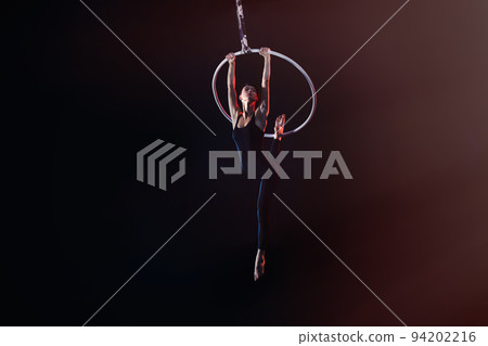 Young woman performing acrobatic element on aerial ring against dark background Young woman performing acrobatic element on aerial ring against dark background 94202216