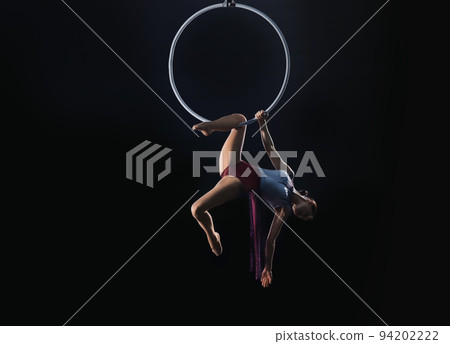 Young woman performing acrobatic element on aerial ring against dark background. Space for text 94202222