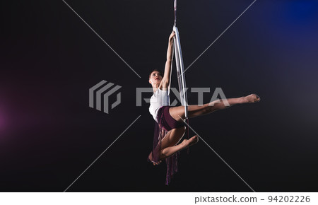 Young woman performing acrobatic element on aerial ring against dark background. Space for text Young woman performing acrobatic element on aerial ring against dark background. Space for text 94202226
