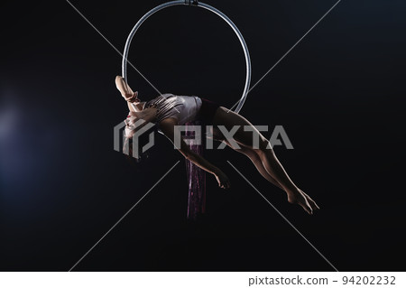 Young woman performing acrobatic element on aerial ring against dark background Young woman performing acrobatic element on aerial ring against dark background 94202232