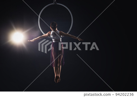 Young woman performing acrobatic element on aerial ring against dark background. Space for text 94202236