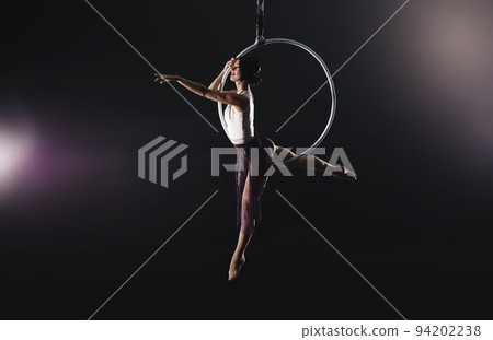 Young woman performing acrobatic element on aerial ring against dark background Young woman performing acrobatic element on aerial ring against dark background 94202238