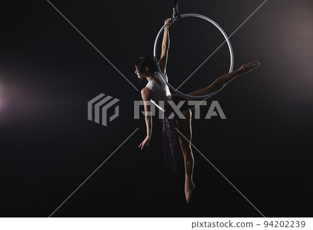 Young woman performing acrobatic element on aerial ring against dark background 94202239